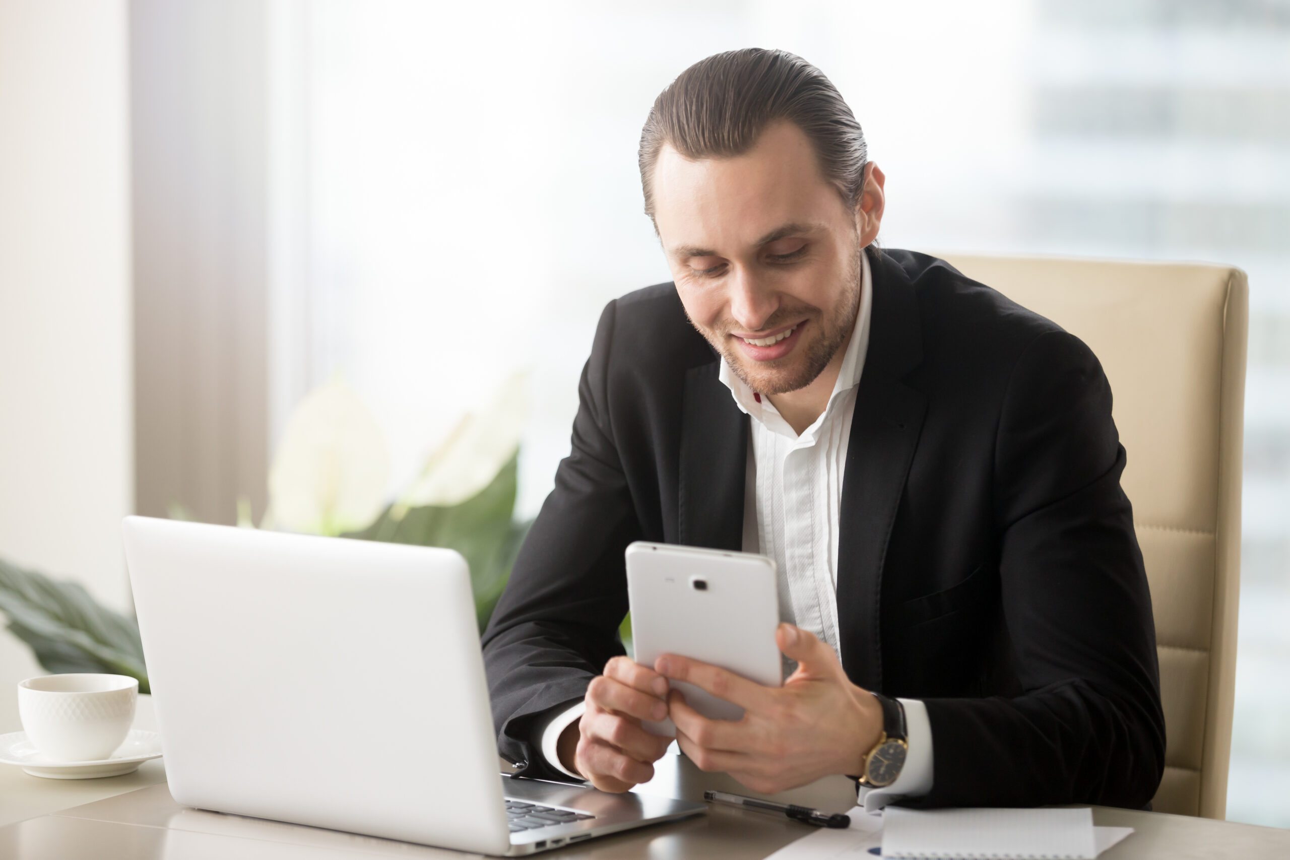 Smart young smiling handsome businessman in modern office in front of laptop looking at electronic tablet. Receiving good news, startup and entrepreneurship, financial success and investing concept.